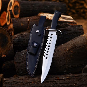 May include: A silver and black hunting knife with a black leather sheath. The knife is resting on a pile of logs.
