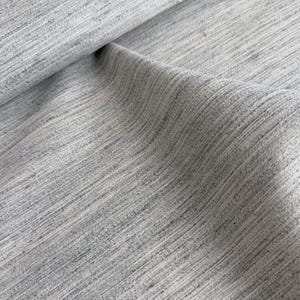 May include: Close-up of a light gray fabric with a textured, woven appearance. The fabric has a subtle, striped pattern created by the weave, with variations in tone. The fabric is folded, showing depth and dimension.