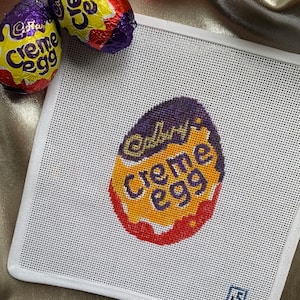 May include: A white canvas with a stitched design of a Cadbury Creme Egg. The egg design is purple, yellow, and red. Two foil-wrapped Cadbury Creme Eggs are in the background.