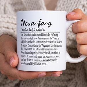 Könnte beinhalten: Weiße Keramik-Tasse mit dem deutschen Wort "Neuanfang" und seiner Definition. Die Tasse wird von einer Person gehalten, die einen cremefarbenen Strickpullover trägt. Das Design der Tasse enthält eine Definition des Wortes.