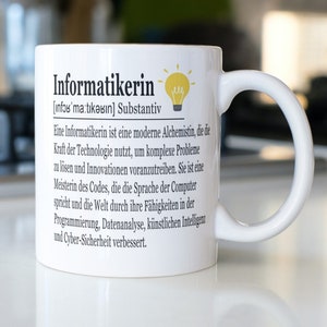 Könnte beinhalten: Weißer Keramikbecher mit dem deutschen Wort "Informatikerin" aufgedruckt, zusammen mit einer Definition und Beschreibung. Eine gelbe Glühbirnen-Grafik ist ebenfalls vorhanden. Der Becher hat einen weißen Henkel und steht auf einer weißen Oberfläche.