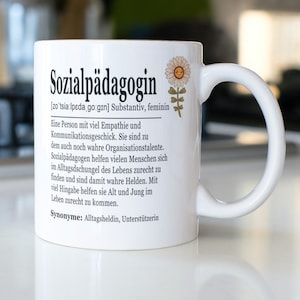 Könnte beinhalten: Eine weiße Keramiktasse mit einer schwarz-weißen Definition des Wortes "Sozialpädagogin" auf Deutsch. Die Definition beinhaltet die Aussprache, die Wortart und eine Beschreibung der Rolle. Eine kleine Blume mit einem Smiley ist auf der Tasse abgebildet.