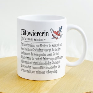 Op de afbeelding: Witte keramische mok met een zwart-wit ontwerp met de tekst "Tätowiererin" en een definitie van het woord. De mok heeft ook een klein rood hart met een dolk erdoorheen en het woord "LOVE" erop.