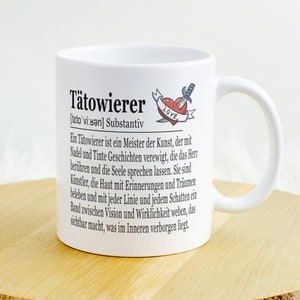 Op de afbeelding: Witte keramische mok met een zwart en rood tattoo-geïnspireerd ontwerp. De mok heeft het woord "Tätowierer" in zwarte tekst met een definitie in het Duits. Het ontwerp bevat ook een rood hart met een dolk erdoorheen en het woord "LOVE" in zwarte tekst.