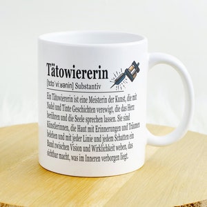 Op de afbeelding: Een witte keramische mok met een zwart-witte illustratie van een tatoeagemachine en de tekst "Tätowiererin [teto'vi sənin] Substantiv Ein Tätowiererin ist eine Meisterin der Kunst, die mit Nadel und Tinte Geschichten verewigt, die das Herz berühren und die Seele sprechen lassen. Sie sind Künstlerinnen, die Haut mit Erinnerungen und Tränen beleben und mit jeder Linie und jedem Schatten ein Band zwischen Vision und Wirklichkeit weben, das sichtbar macht, was im Inneren verborgen liegt."