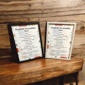 Könnte beinhalten: Zwei gerahmte Drucke mit dem deutschen Text "Rezept für ein perfektes Team". Ein Rahmen ist schwarz, der andere weiß. Die Drucke haben ein Blumendesign und werden auf einer Holzoberfläche ausgestellt.