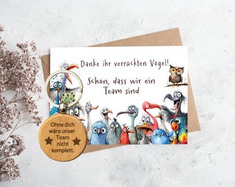 May include: A wooden keychain with a message that reads "Ohne dich wäre unser Team nicht komplett."  The keychain features a cartoon illustration of a group of birds with the text "Danke ihr verrückten Vögel!" and "Schön, dass wir ein Team sind".