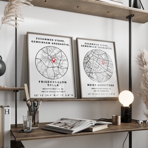 May include: Two black and white framed prints with a map of a city and the text "Zusammen Stark, Gemeinsam Grobartig."  The prints are hanging on a wall above a wooden desk with a book, a glass of water, a pencil holder, and a black and white lamp.