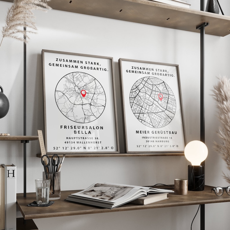 May include: Two black and white framed prints with a map of a city and the text "Zusammen Stark, Gemeinsam Grobartig."  The prints are hanging on a wall above a wooden desk with a book, a glass of water, a pencil holder, and a black and white lamp.