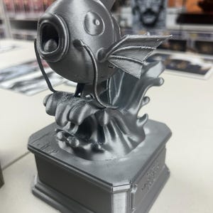 May include: A metallic gray 3D printed figurine of a fish character with a wide open mouth, long whiskers, and a dorsal fin, mounted on a rectangular base. The base features embossed text on the front panel, displaying the name Magikarp.