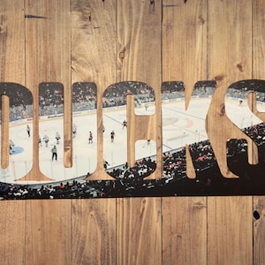May include: A wooden sign with the word "DUCKS" cut out, revealing a hockey game scene. The sign is made of dark metal and mounted on a wooden background with vertical planks.