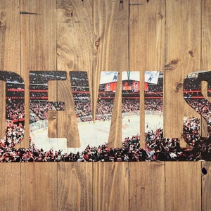 May include: A wooden sign with the word "DEVILS" spelled out in large letters. The letters are cut out and the background is a photo of a hockey arena filled with fans.