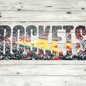 May include: The word "ROCKETS" in large, textured letters, filled with a colorful image of a basketball arena, set against a weathered, white-washed wood background. The letters are dark with a textured appearance.