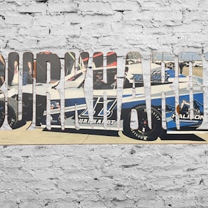 May include: A wooden sign with the word "BURKART" spelled out in large, black letters. The letters are cut out and the background is a photo of a race car with the number 27 on the side. The car is blue and white with black accents.