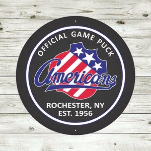 May include: A round, black sign with a white border featuring the text "OFFICIAL GAME PUCK" and "Americans" in a stylized script. The sign includes a shield design with red and white stripes and three stars, along with "ROCHESTER, NY EST. 1956".