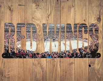 Op de afbeelding: Een metalen bord met het woord "RED WINGS" uitgesneden. De achtergrond is een foto van een hockeyarena met fans op de tribunes.