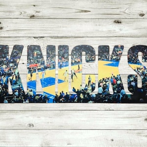May include: Metal sign featuring the word "KNICKS" in large, bold letters. The letters are filled with a collage of basketball game action, including players and spectators. The sign is mounted on a white wooden background.