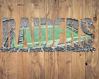 May include: A metal sign with the word "RAIDERS" cut out, revealing a view of a football stadium with a green pitch and a crowd of people. The stadium has the words "LAS VEGAS" on the pitch.