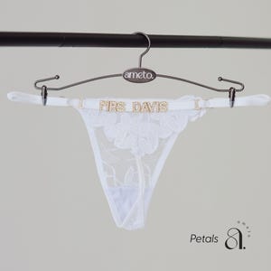 May include: White lace thong underwear with floral embroidery, displayed on a hanger. The hanger has the brand name "ameto" and the text "MRS DAVIS" in gold lettering. The underwear is suspended from a metal hanger.