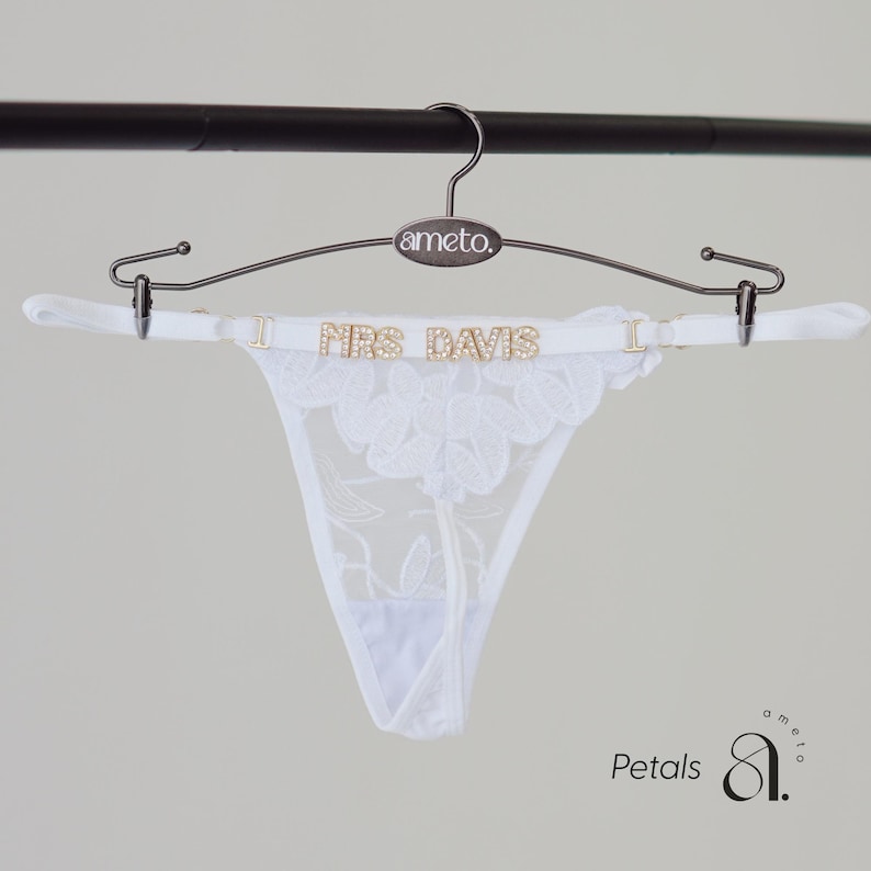 May include: White lace thong underwear with floral embroidery, displayed on a hanger. The hanger has the brand name "ameto" and the text "MRS DAVIS" in gold lettering. The underwear is suspended from a metal hanger.