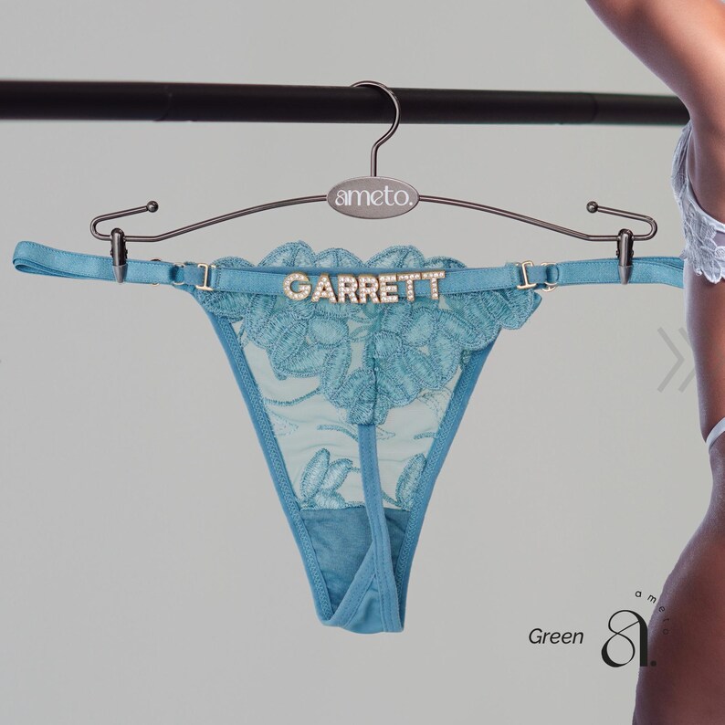 May include: A teal thong with floral lace detailing and the name "GARRETT" in silver letters. The thong is displayed on a hanger with the brand name "ameto". The word "Green" is visible in the bottom right corner.