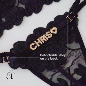 May include: Black lingerie set with floral lace detailing. The set includes a thong with a detachable strap and a decorative gold-toned nameplate that reads "CHRIS" with a heart accent. The lingerie is designed with adjustable straps.