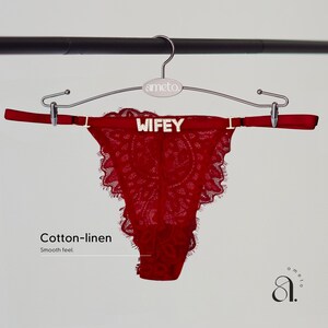 Pode incluir: Uma tanga de renda vermelha com a inscri&ccedil;&atilde;o "WIFEY" na cintura, pendurada em um cabide prateado. A tanga tem uma borda ondulada e &eacute; feita de algod&atilde;o e linho. O cabide tem a marca "ameto".