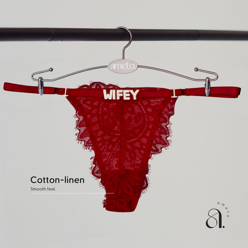 Pode incluir: Uma tanga de renda vermelha com a inscri&ccedil;&atilde;o "WIFEY" na cintura, pendurada em um cabide prateado. A tanga tem uma borda ondulada e &eacute; feita de algod&atilde;o e linho. O cabide tem a marca "ameto".
