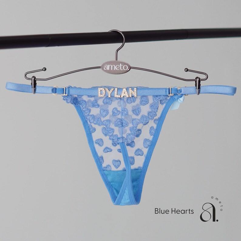 May include: Light blue thong underwear with a heart pattern, hanging from a silver hanger. The word "DYLAN" is spelled out in rhinestones across the waistband. The words "Blue Hearts" are printed in blue text.