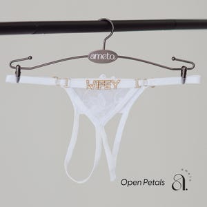 May include: White lingerie set with the word "WIFEY" spelled out in gold letters. The set is displayed on a metal hanger with the brand name "ameto." The image also includes the text "Open Petals."