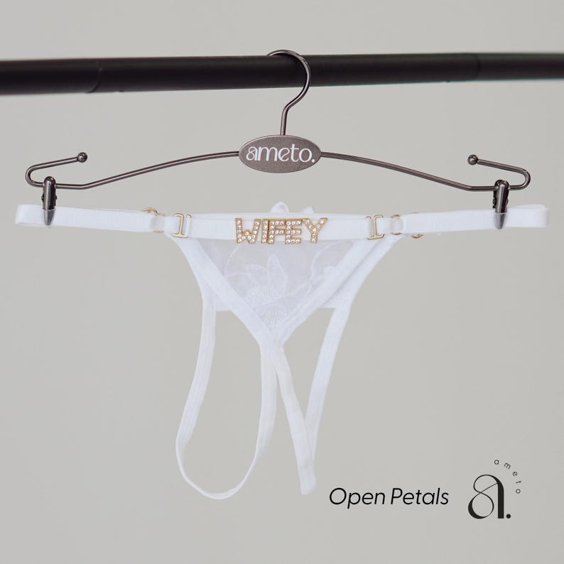 May include: White lingerie set with the word "WIFEY" spelled out in gold letters. The set is displayed on a metal hanger with the brand name "ameto." The image also includes the text "Open Petals."