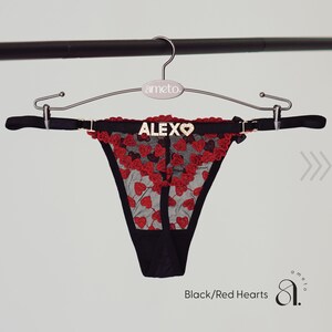 May include: Black thong underwear with red heart embroidery and trim, hanging on a silver hanger. The word "ALEX" is spelled out in silver letters on the waistband. The text "Black/Red Hearts" is visible at the bottom.
