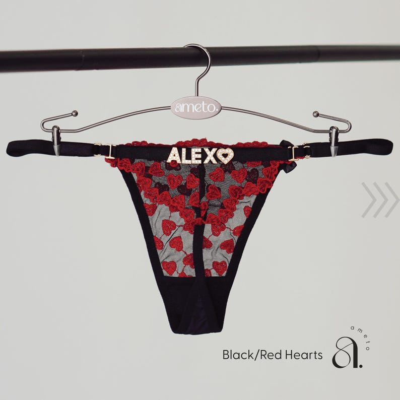 May include: Black thong underwear with red heart embroidery and trim, hanging on a silver hanger. The word "ALEX" is spelled out in silver letters on the waistband. The text "Black/Red Hearts" is visible at the bottom.
