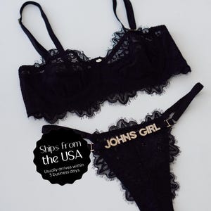 May include: Black lace lingerie set featuring a bra and thong. The bra has adjustable straps and delicate lace trim. The thong has the text "JOHNS GIRL" in sparkling letters across the waistband. The set is displayed on a white background.