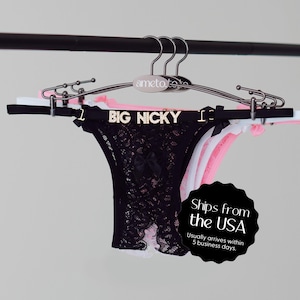 May include: A display of several thong underwear in various colors, including black, white, and pink, hanging on a metal hanger. The black thong features a lace design and a small bow. The text "BIG NICKY" is displayed on the black thong.