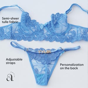 May include: Light blue lingerie set featuring a semi-sheer tulle fabric. The set includes a bra and a thong with adjustable straps. The thong has the name "TYLER" on the front. The bra has a bow in the center. The lingerie is designed with floral lace details.