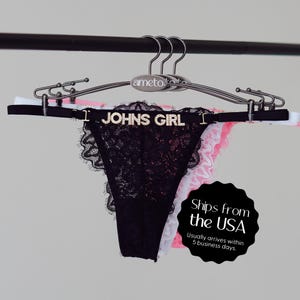 Pode incluir: Calcinha tanga de renda preta com o texto "JOHNS GIRL" em letras prateadas. A roupa &iacute;ntima est&aacute; pendurada em um cabide prateado com roupa &iacute;ntima branca, rosa e preta atr&aacute;s.