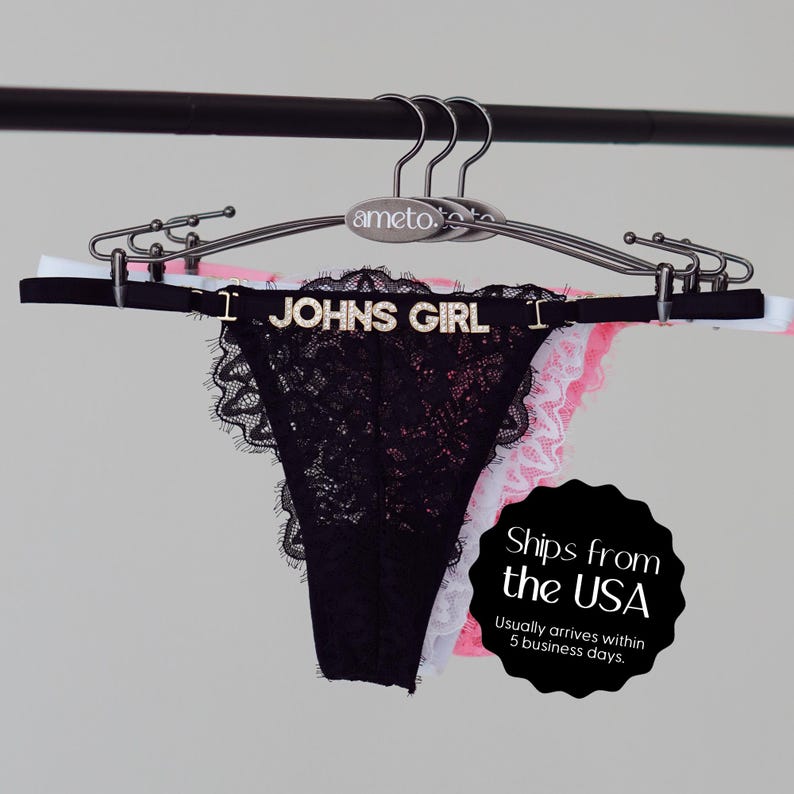 Pode incluir: Calcinha tanga de renda preta com o texto "JOHNS GIRL" em letras prateadas. A roupa &iacute;ntima est&aacute; pendurada em um cabide prateado com roupa &iacute;ntima branca, rosa e preta atr&aacute;s.