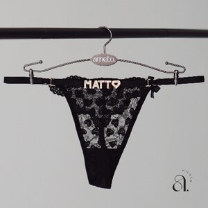 以下が含まれることがあります： 黒いレースのTバックショーツで、ウエストバンドに「MATTO」の文字が金色で入っています。下着は「ameto」のブランド名がプリントされたシルバーのハンガーに吊るされています。小さな黒いリボンが側面にあります。
