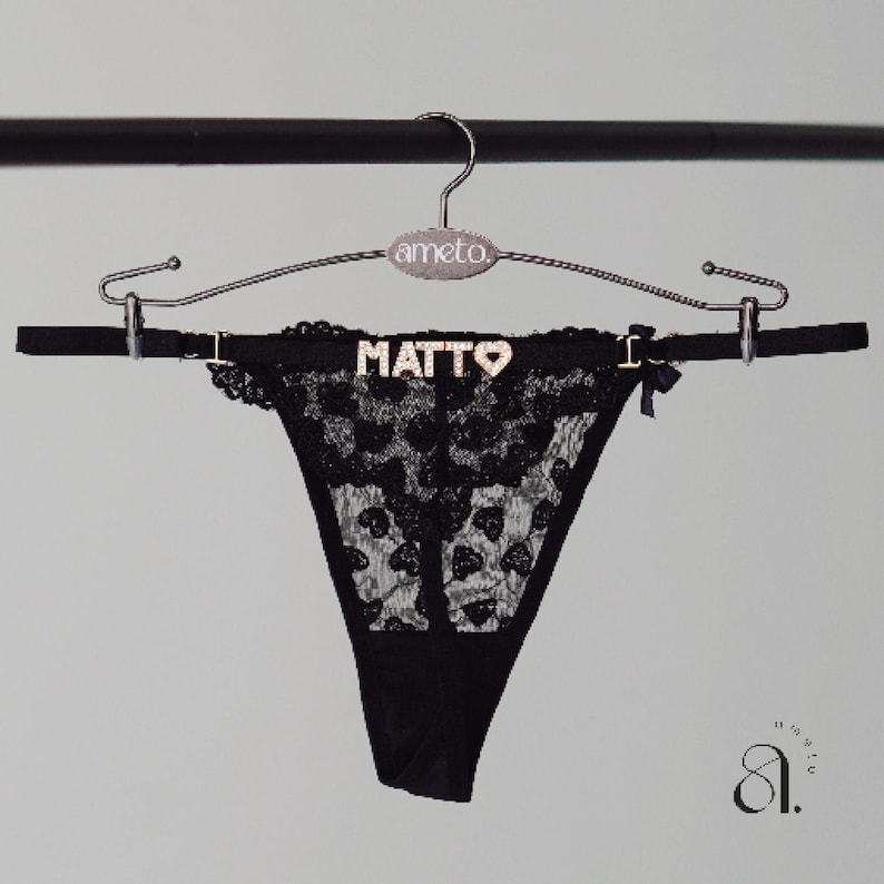 以下が含まれることがあります： 黒いレースのTバックショーツで、ウエストバンドに「MATTO」の文字が金色で入っています。下着は「ameto」のブランド名がプリントされたシルバーのハンガーに吊るされています。小さな黒いリボンが側面にあります。