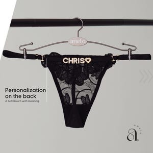 May include: Black thong underwear with floral lace detailing, displayed on a hanger. The word "CHRIS" is spelled out in rhinestones across the back, with a heart symbol. The hanger is silver and has the brand name "ameto" on it.