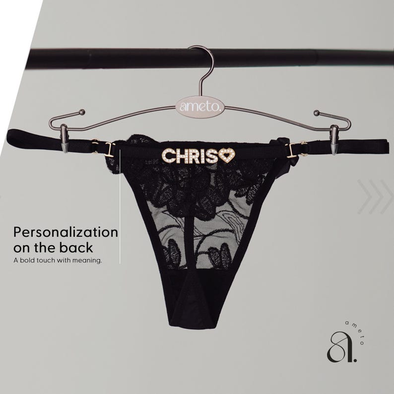 May include: Black thong underwear with floral lace detailing, displayed on a hanger. The word "CHRIS" is spelled out in rhinestones across the back, with a heart symbol. The hanger is silver and has the brand name "ameto" on it.