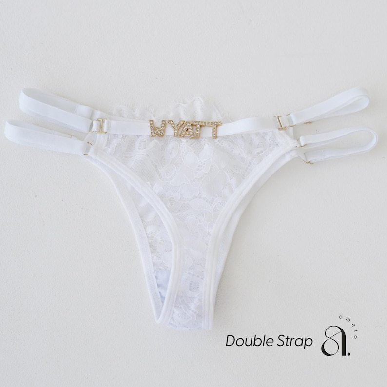 May include: White lace thong with double straps and gold-tone hardware. The word "WYATT" is spelled out in gold-colored rhinestones across the front. The thong features a delicate floral lace pattern.