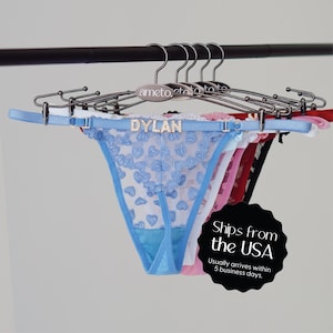 May include: A collection of thong underwear in various colors, including blue, pink, and red, hanging on a rack. The blue thong features a heart pattern and the name "DYLAN" in rhinestones.