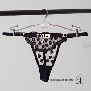 May include: Black thong underwear with a heart-shaped lace design. The underwear is displayed on a hanger with the name "TYLER" in gold letters. The text "Black/Nude Hearts" is visible.