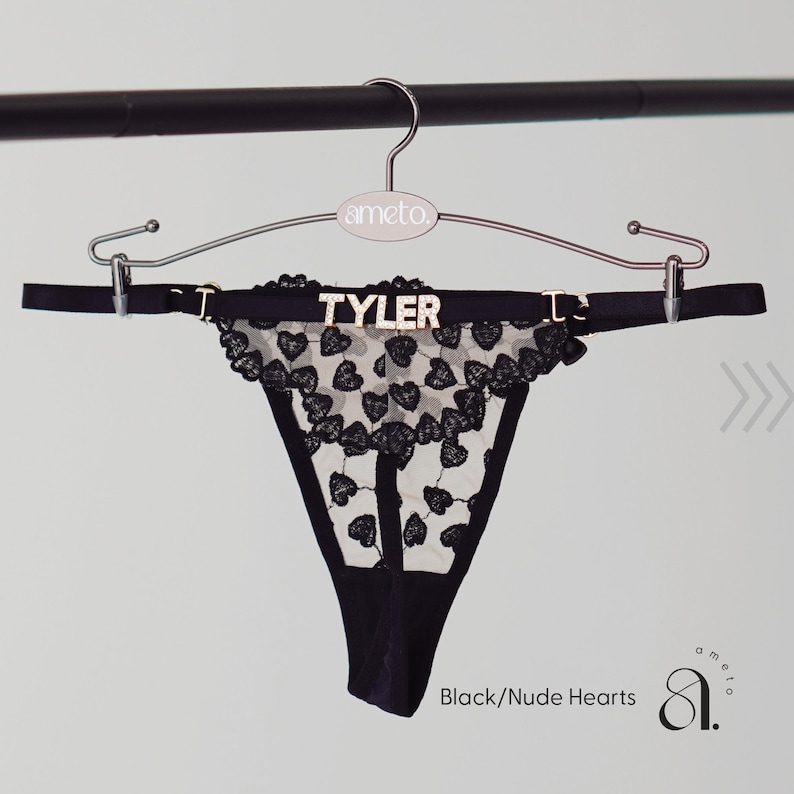 May include: Black thong underwear with a heart-shaped lace design. The underwear is displayed on a hanger with the name "TYLER" in gold letters. The text "Black/Nude Hearts" is visible.