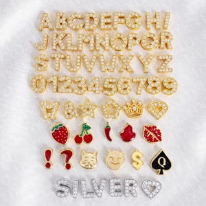 May include: A collection of gold-colored alphabet letters, numbers, and various charms, all encrusted with small, sparkling stones. The charms include a strawberry, cherries, a chili pepper, and a cat face.