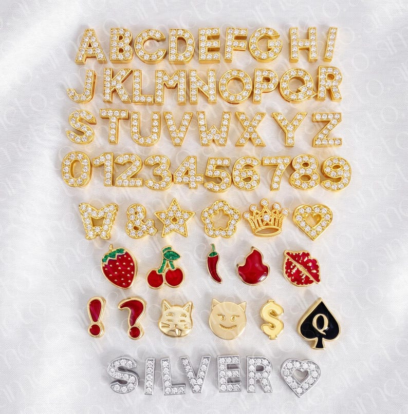 May include: A collection of gold-colored alphabet letters, numbers, and various charms, all encrusted with small, sparkling stones. The charms include a strawberry, cherries, a chili pepper, and a cat face.
