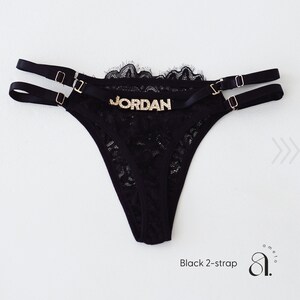 Pode incluir: Tanga de renda preta com duas al&ccedil;as e ferragens douradas. A palavra "JORDAN" est&aacute; escrita em letras douradas na frente. A borda superior &eacute; adornada com renda preta. O texto "Black 2-strap" est&aacute; na parte inferior.