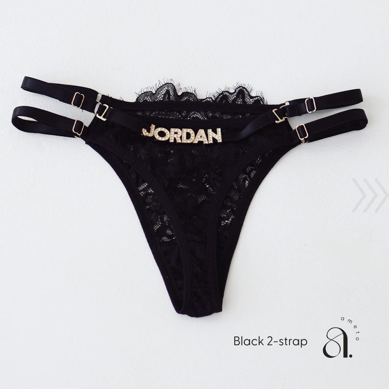 Pode incluir: Tanga de renda preta com duas al&ccedil;as e ferragens douradas. A palavra "JORDAN" est&aacute; escrita em letras douradas na frente. A borda superior &eacute; adornada com renda preta. O texto "Black 2-strap" est&aacute; na parte inferior.